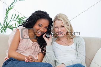 Two women listening to a mobile phone are smiling at the camera