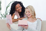 A woman opens a box containing a present and her friend smiles
