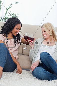 Two women are sitting on the ground talking and holding wine gla