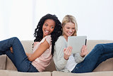 Two women sitting back to back on a couch looking at a tablet co