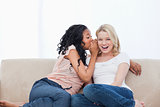 A woman sitting on a couch is whispering into her friends ear