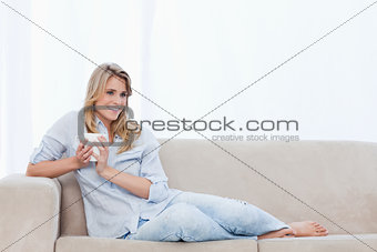 A smiling woman lying on a couch is holding a cup of coffee