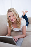 A woman smiling at the camera is lying on a couch with a laptop 