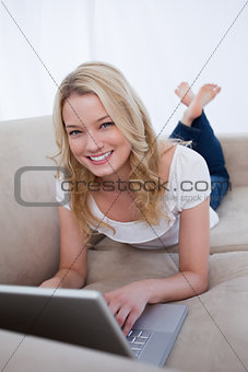 A woman smiling at the camera is lying on a couch with a laptop 