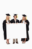 Three students in graduate robe holding a blank sign
