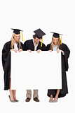 Three smiling students in graduate robe holding a blank sign
