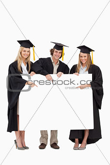 Three students in graduate robe holding and pointing a blank sig