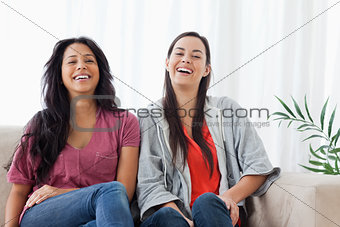 Two women sit beside on another on the couch while laughing 