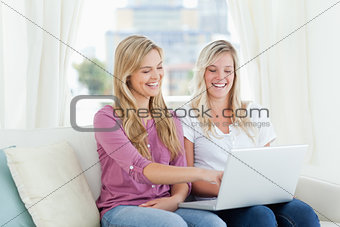 A pair of laughing women sitting on the couch with a laptop in h