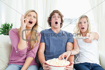 Three friends enjoying popcorn together while shocked at the tv 
