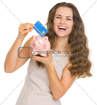 Happy young woman putting credit card into piggy bank