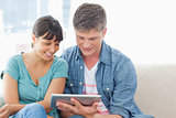 A smiling couple sit on the couch and use a tablet 