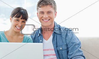 A couple with a laptop sitting as they smile and look into the c