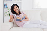 Smiling woman holding a book and a mug while lying on a couch