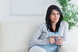 Thinking woman sits on a couch while holding a mug
