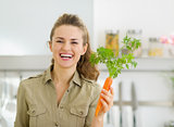 Smiling young housewife holding carrot in kitchen
