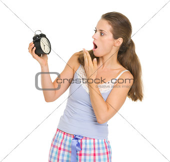 Shocked young woman in pajamas looking on alarm clock