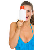 Happy young woman in swimsuit hiding behind sun screen creme