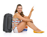 Happy young tourist woman sitting near wheel bag and pointing on