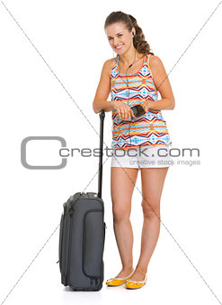 Smiling young tourist woman with wheel bag listening music