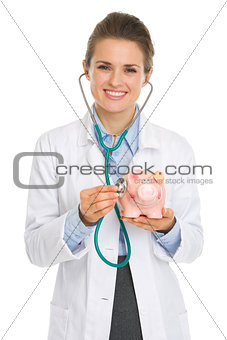 Smiling medical doctor woman listening piggy bank with stethosco