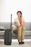 Happy young woman in living room with travel bag talking mobile 
