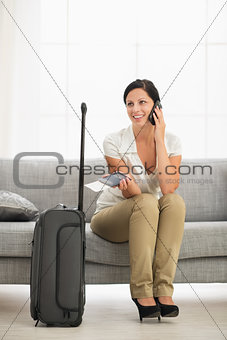 Happy young woman in living room with travel bag talking mobile 