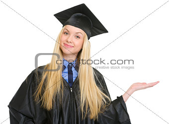 Happy young woman in graduation gown presenting something on emp