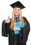 Happy young woman in graduation gown with books showing thumbs u