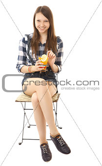Girl teenager, caucasian appearance, brunette, wearing a plaid shirt and short denim shorts, holding a glass of drink. Girl is relaxing, sitting on a folding chair with orange drink in hand.