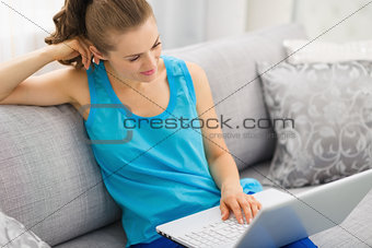 Thoughtful young woman sitting on sofa and using laptop in livin
