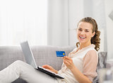 Happy young housewife laying on sofa with laptop and credit card