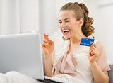 Happy young housewife with laptop and credit card crossed finger