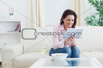 Woman with wine and bowl using digital tablet on sofa