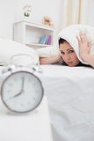 Woman covers ears with sheet as alarm clock rings