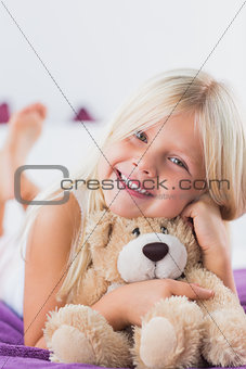 Smiling girl with her teddy bear lying on a bed