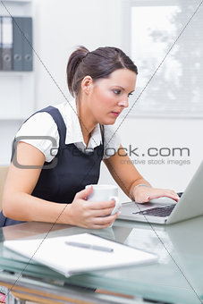 Female executive using laptop as she holds coffee cup at desk
