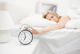 Irritated young woman in bed extending hand to alarm clock