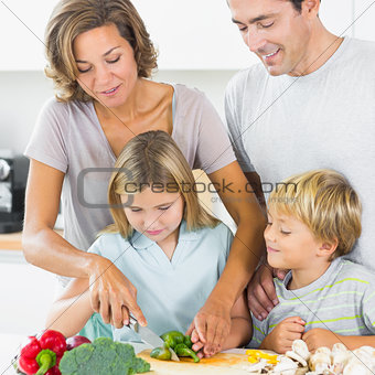 Mother teaching daughter to slice vegetables as father and son are watching