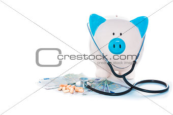 Piggy bank sitting on pile of dollars with stethoscope and pills