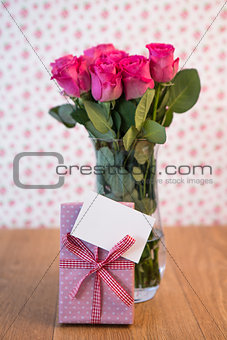 Bunch of pink roses in vase with pink gift leaning against it and blank card