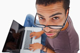 Man wearing glasses sitting on floor using laptop and looking up at camera