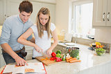 Woman cutting vegetables with man reading the cookbook