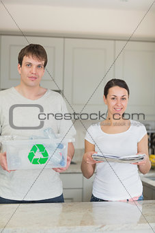 Smiling couple holding newspapers and recycling bin