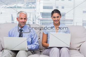 Business people sitting on sofa using their laptops and smiling at camera