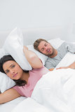 Upset woman covering ears with pillow next to husband snoring