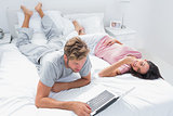 Man using a laptop next to his wife lying on bed