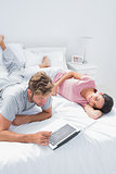 Woman lying on bed while her husband is using a laptop