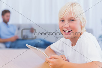 Smiling boy using tablet while his father is reading on the couch