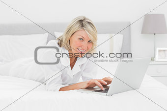 Smiling woman using her laptop on her bed looking at camera